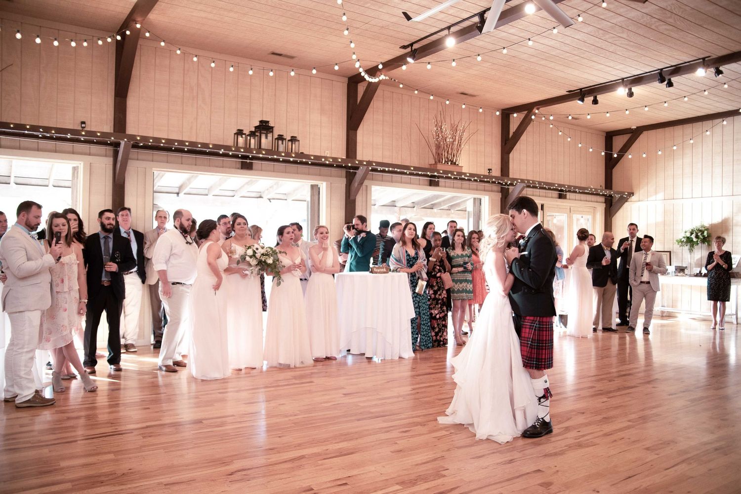 Bride and groom share first dance in rustic barn wedding venue surrounded by bridesmaids and guests.