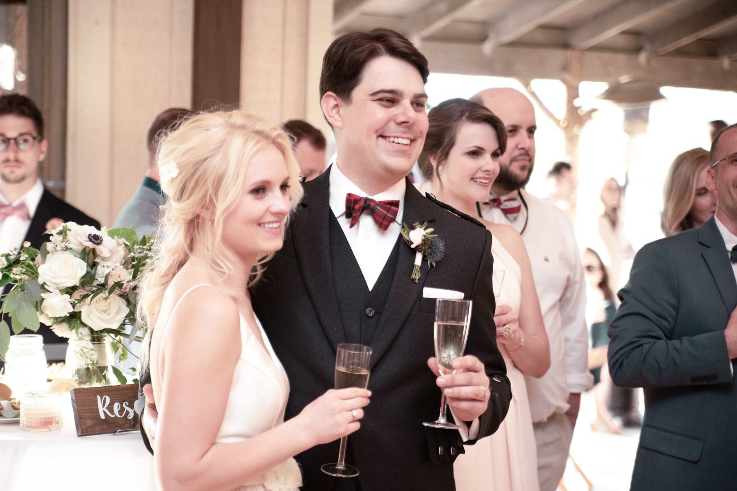 Bride and groom smiling and holding champagne glasses during wedding reception toast with guests in background.