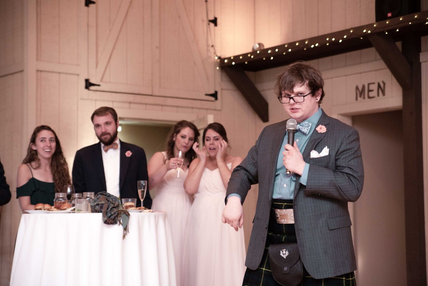 Man in kilt gives wedding speech with microphone while bridesmaids laugh and cry behind him.