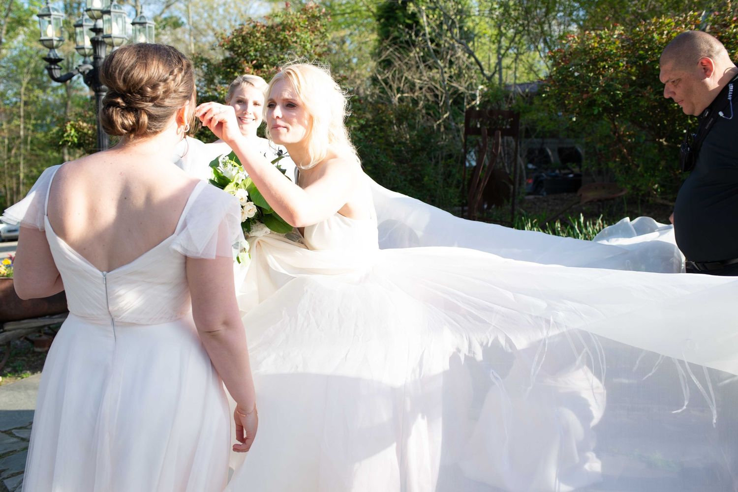 Two brides in white gowns share a joyful moment as a long veil billows behind them at an outdoor wedding ceremony.