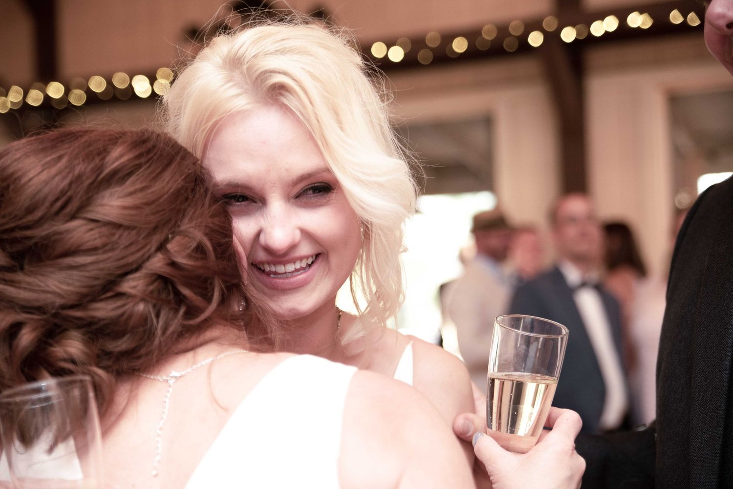 Smiling blonde bride holding champagne glass receives a hug at elegant wedding reception with warm bokeh lights.
