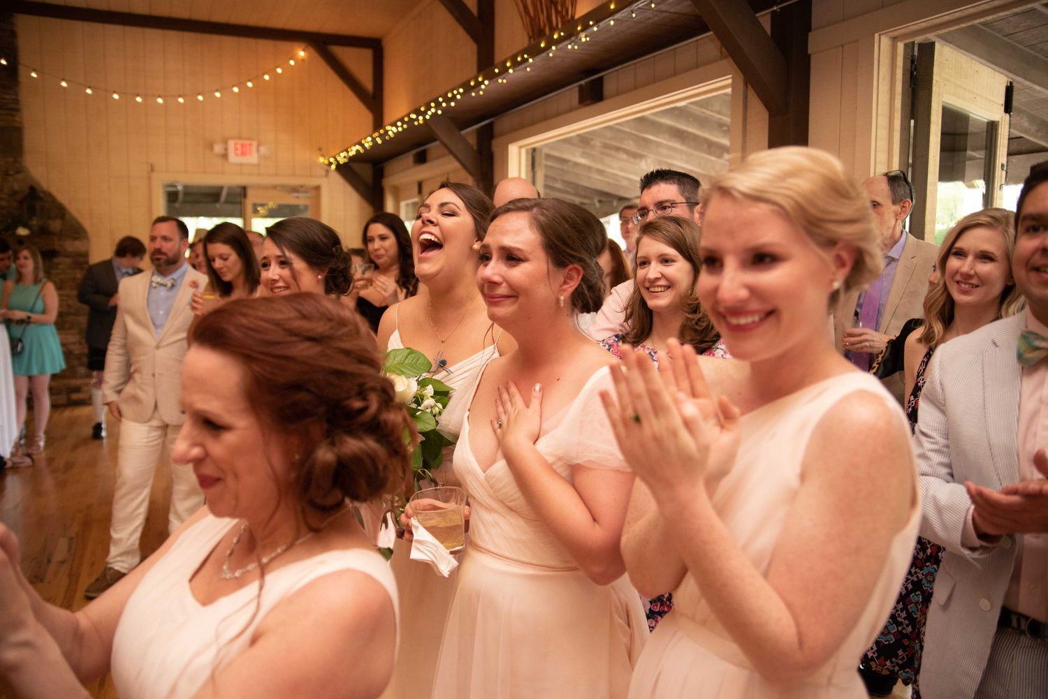 Bridesmaids in blush pink dresses laughing and clapping joyfully at an elegant indoor wedding reception.