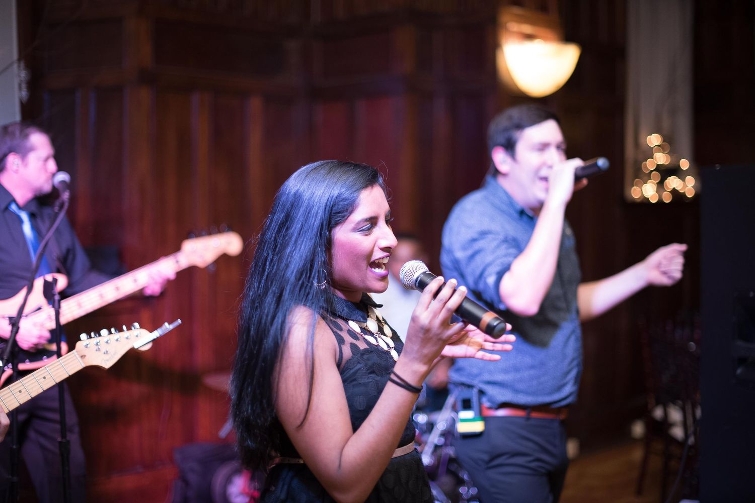 Live band performance with female vocalist singing into microphone, guitarist and male singer performing on stage.