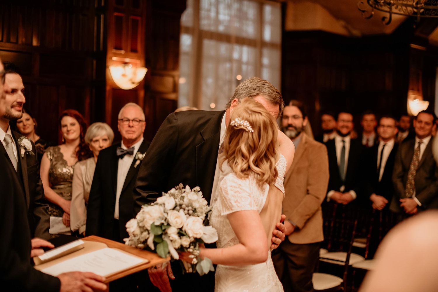 Bride and groom share first kiss at wedding ceremony, bride holding white floral bouquet, guests watching in elegant wood-paneled venue.