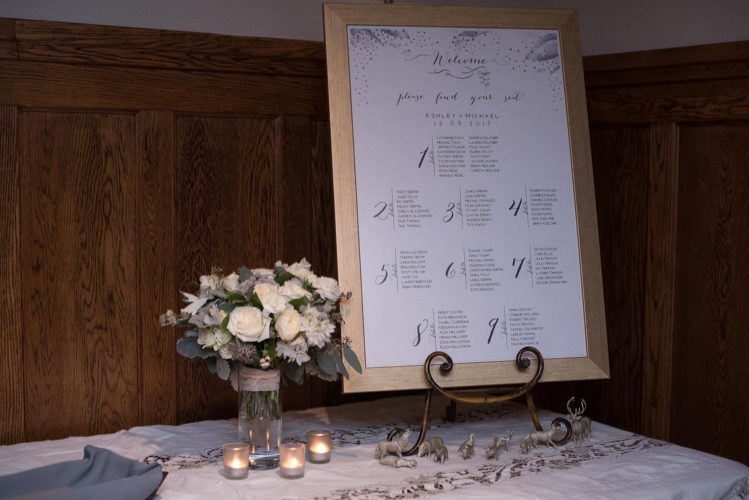Elegant wedding seating chart in gold frame on easel with white floral arrangement and candles on linen-covered table.