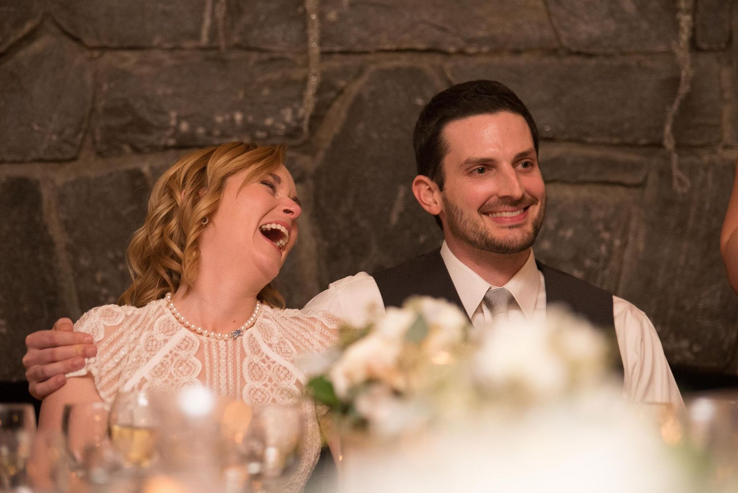 A joyful couple laughing together at a wedding reception, seated at a decorated table in a stone-walled venue.