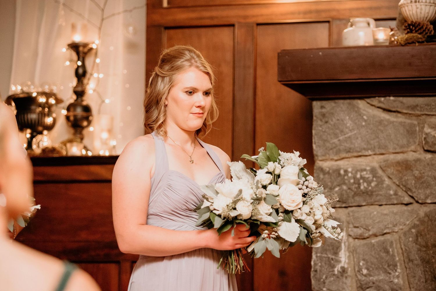 Bridesmaid in gray gown holding white floral bouquet near stone fireplace at elegant wedding venue.