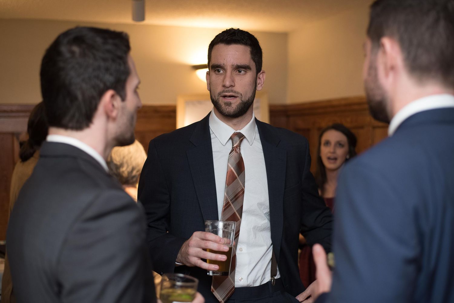 A man in a dark suit holds a drink while conversing with guests at a formal event.