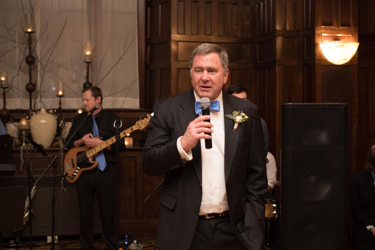 Man in tuxedo gives speech at wedding reception with live saxophonist performing in background.