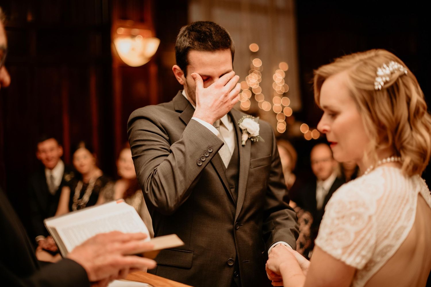 Emotional groom wiping tears during wedding ceremony while bride stands beside him in elegant white dress.