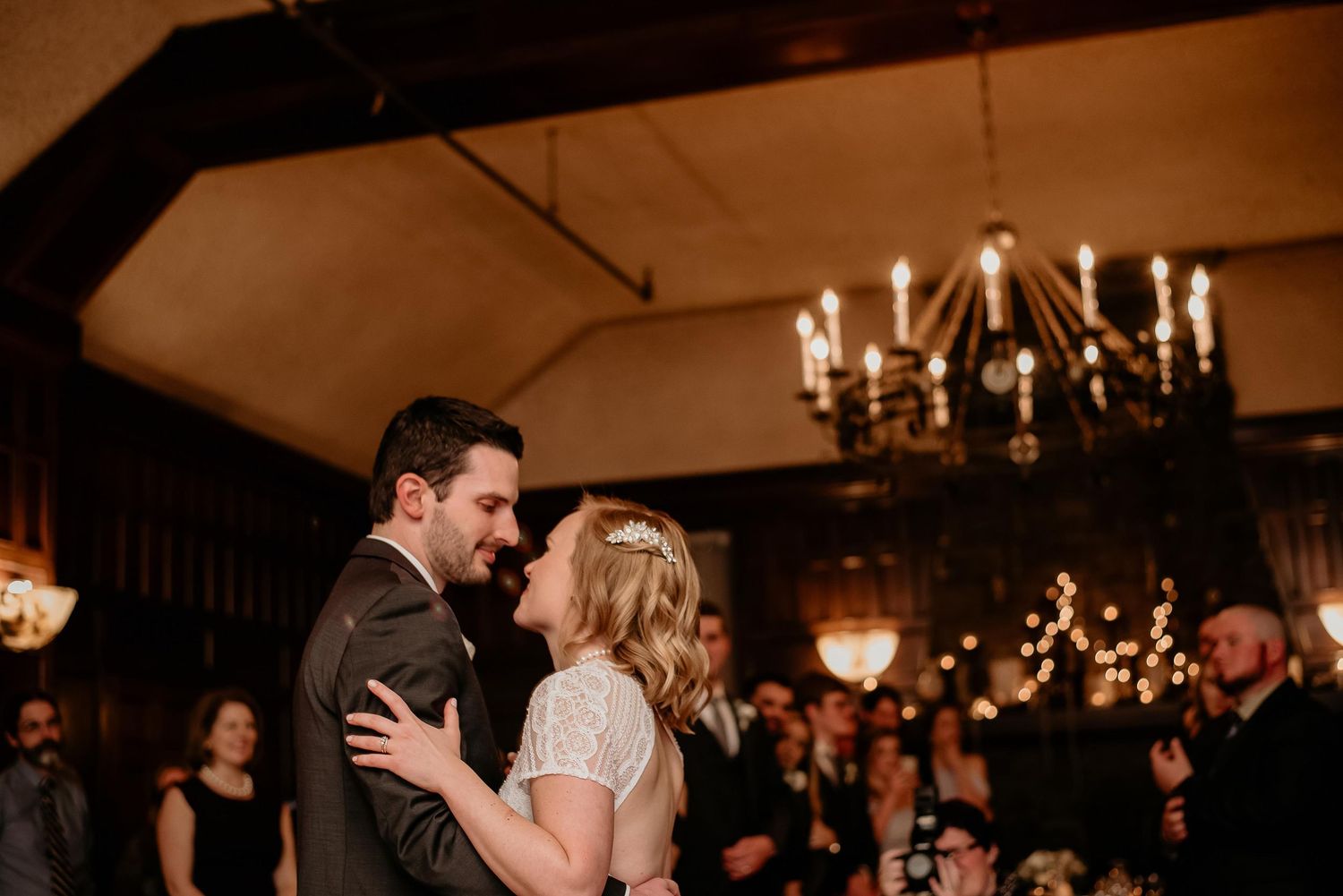 Couple shares their first dance under a chandelier at an elegant wedding reception with guests watching in the background.