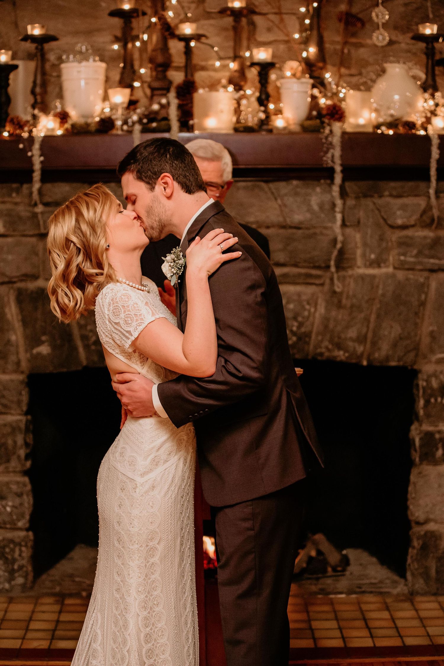 Bride and groom share a romantic kiss in front of a candlelit stone fireplace at their wedding reception.