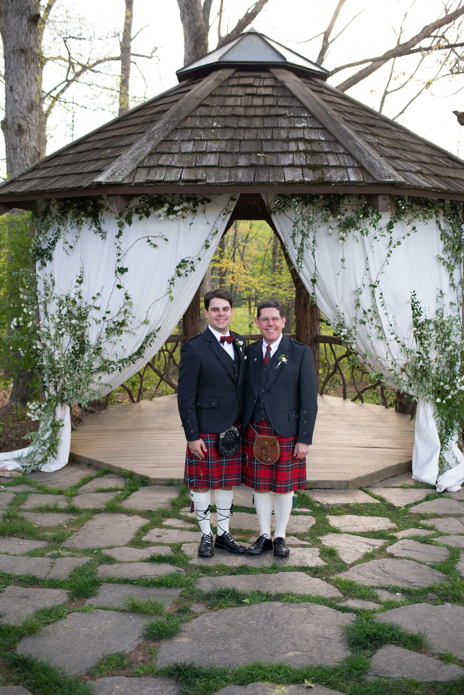 Two grooms in kilts and tuxedo jackets pose before a floral gazebo at an outdoor wedding ceremony.