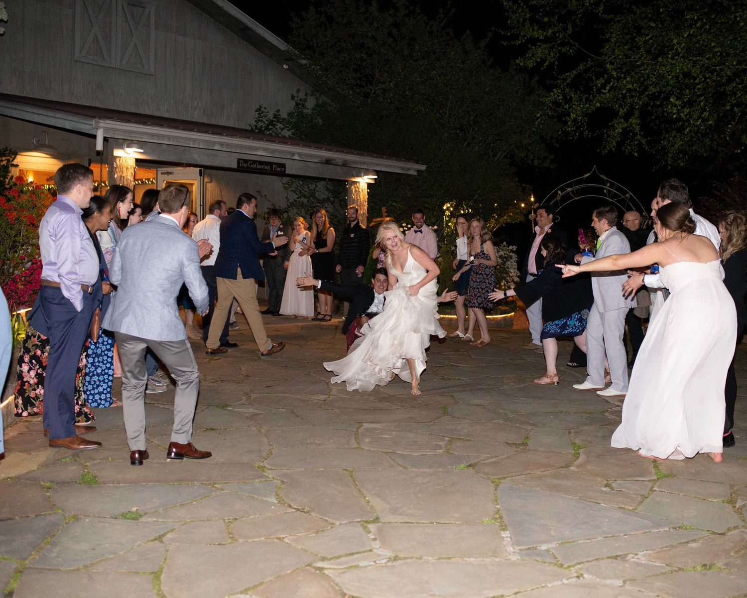 Guests dance outdoors at a night wedding reception on a stone patio surrounded by trees and string lights.