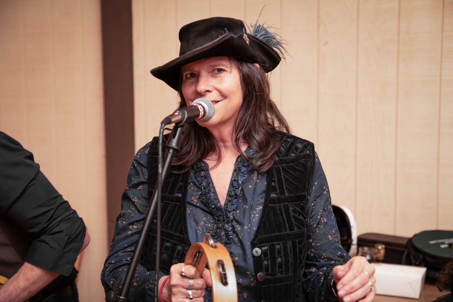 Woman in black feathered hat singing into microphone while holding a ukulele at an indoor music performance.