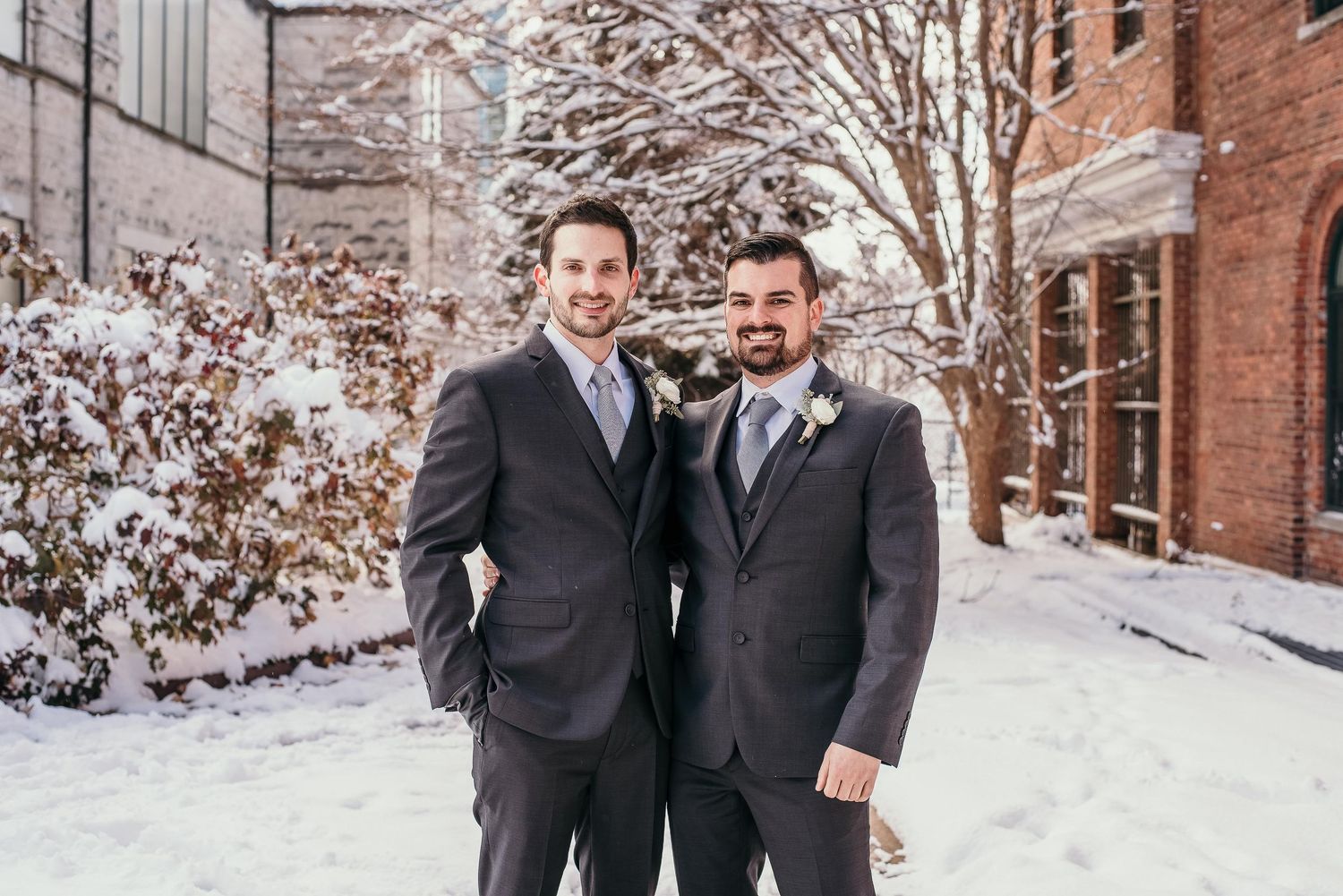 Two grooms in dark suits pose together outdoors in a snowy winter wedding setting near a brick building.