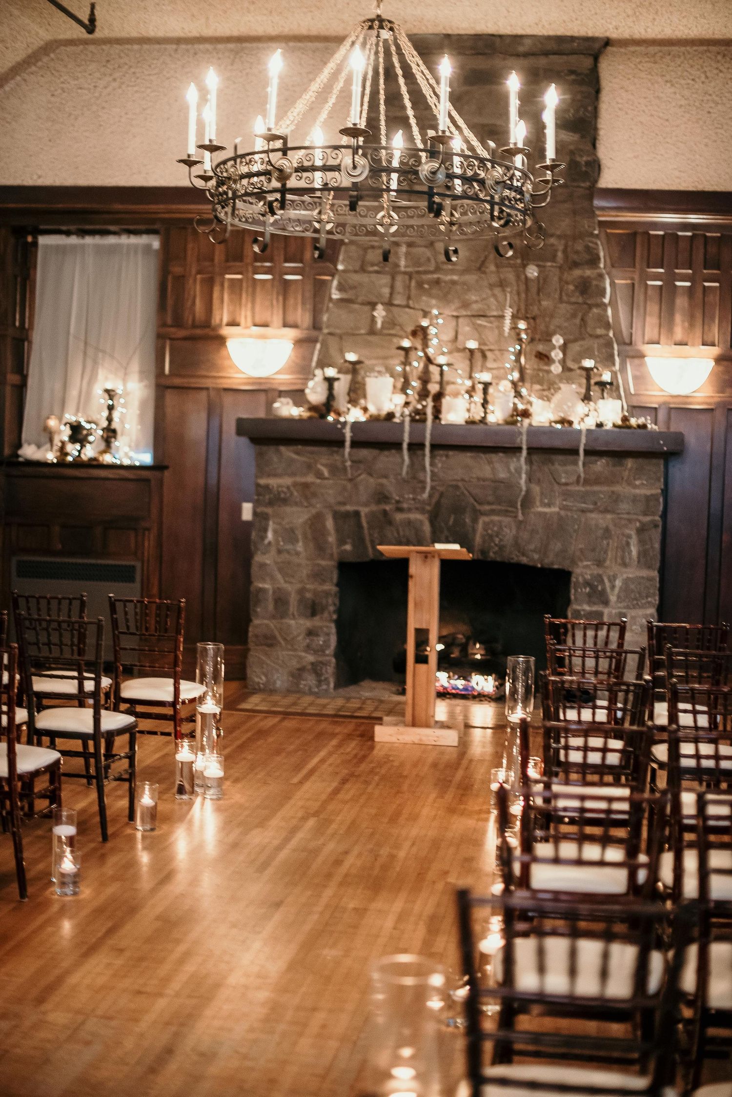 Elegant wedding ceremony setup with chiavari chairs, candles, stone fireplace, and wrought iron chandelier in a rustic venue.