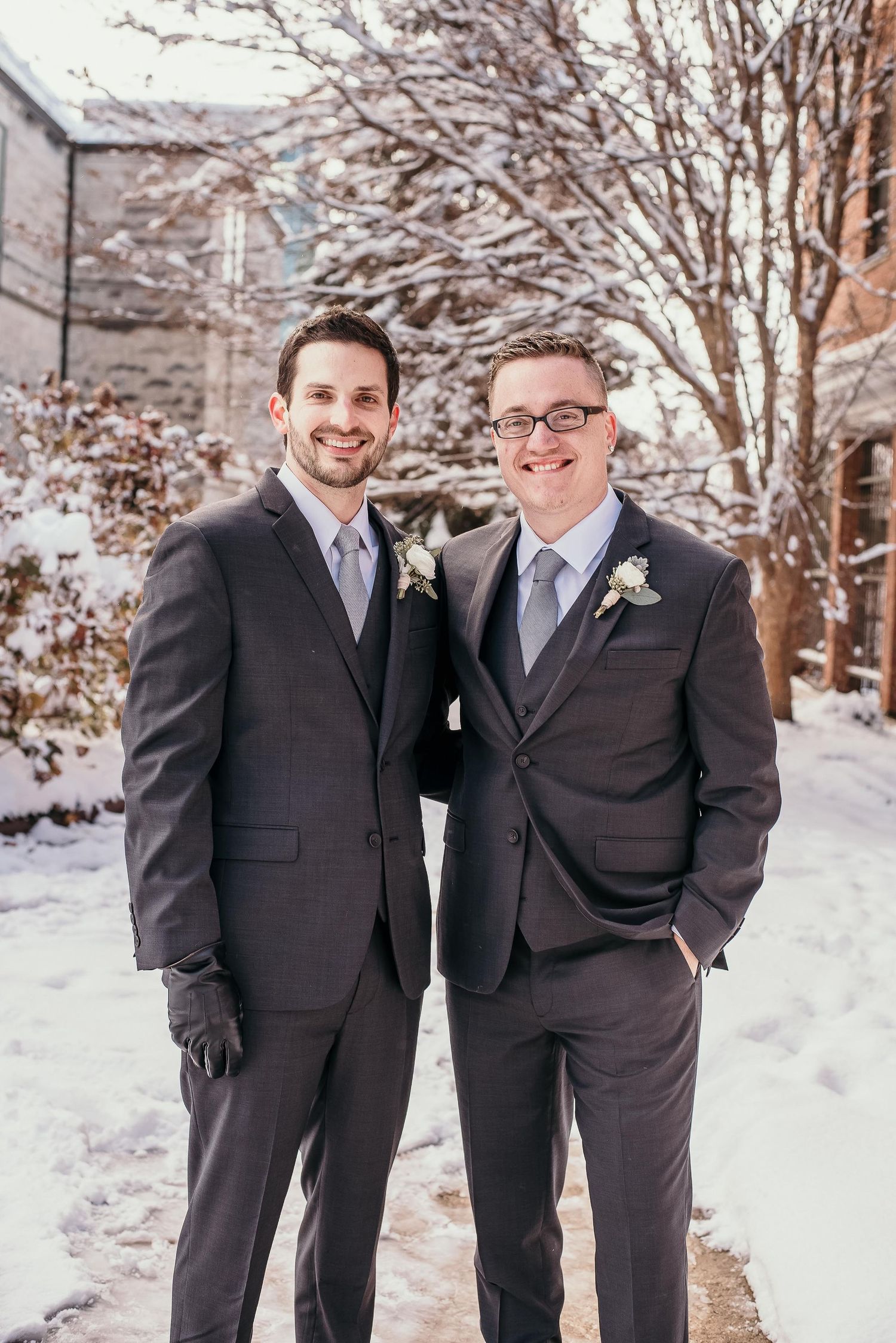 Two grooms in matching dark suits with blue ties pose outdoors in a snowy winter setting on their wedding day.