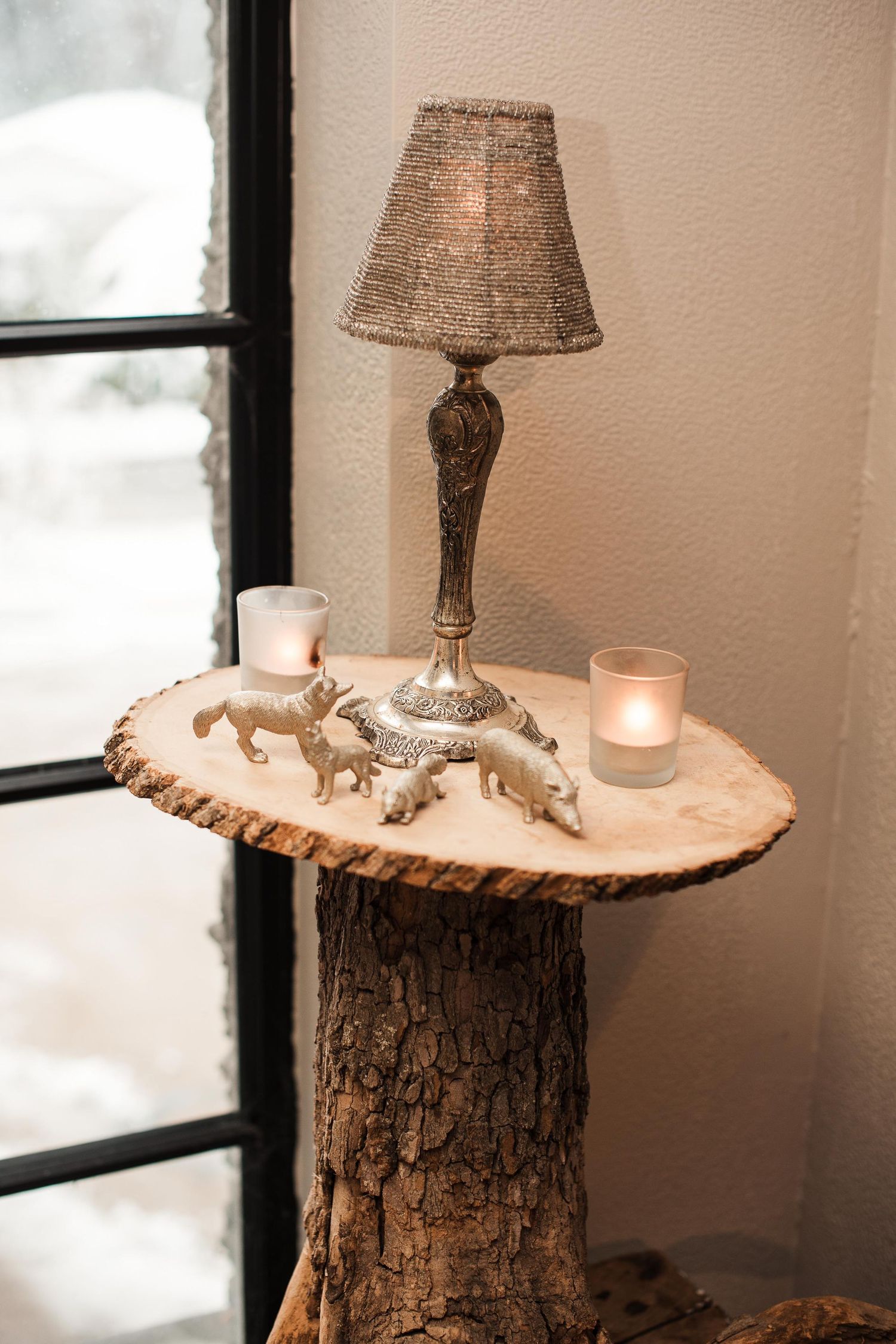 Rustic tree stump side table with small lamp, candles, and silver animal figurines near a snowy window.