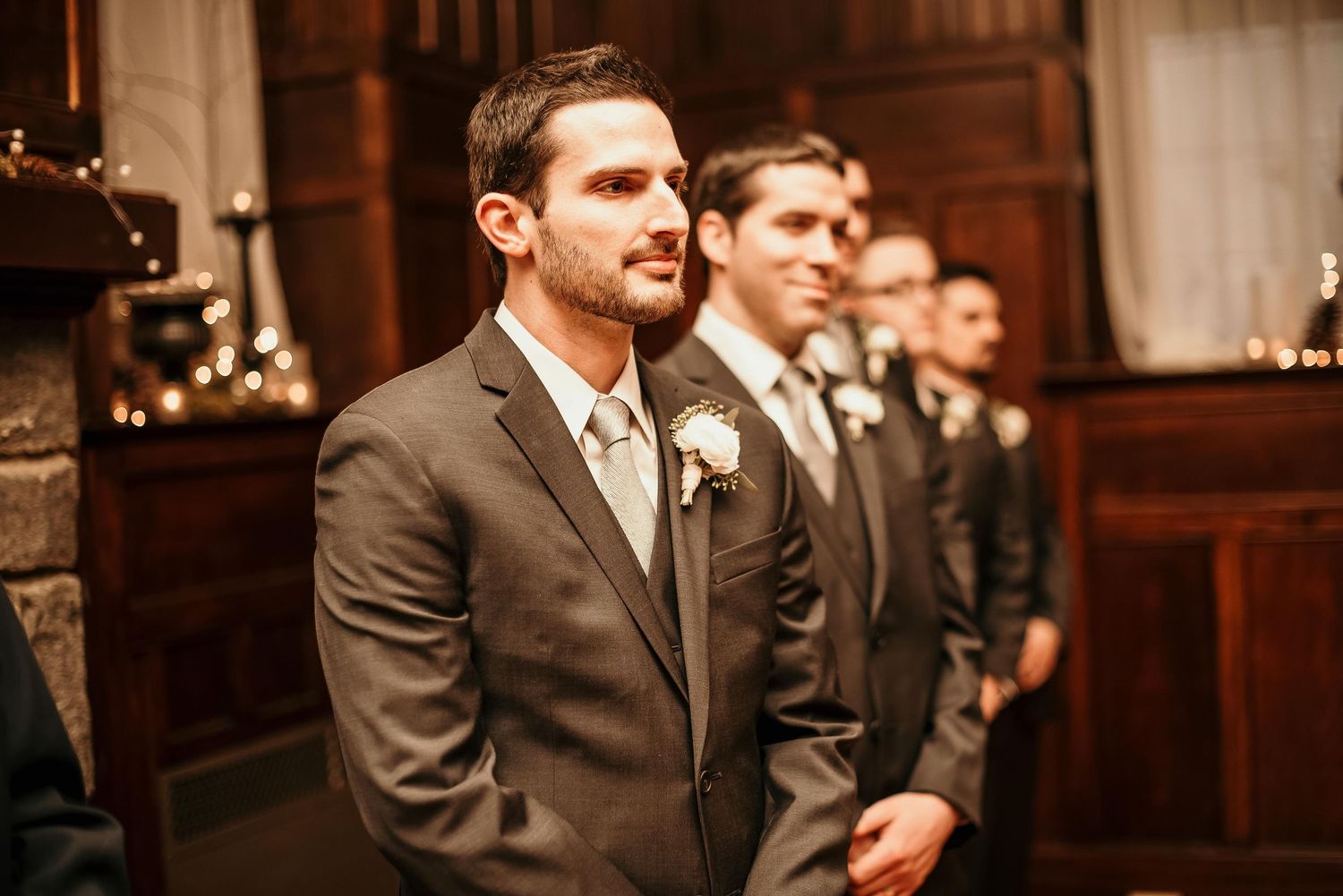 Groom in gray suit with boutonniere stands at altar with groomsmen during wedding ceremony in elegant venue.