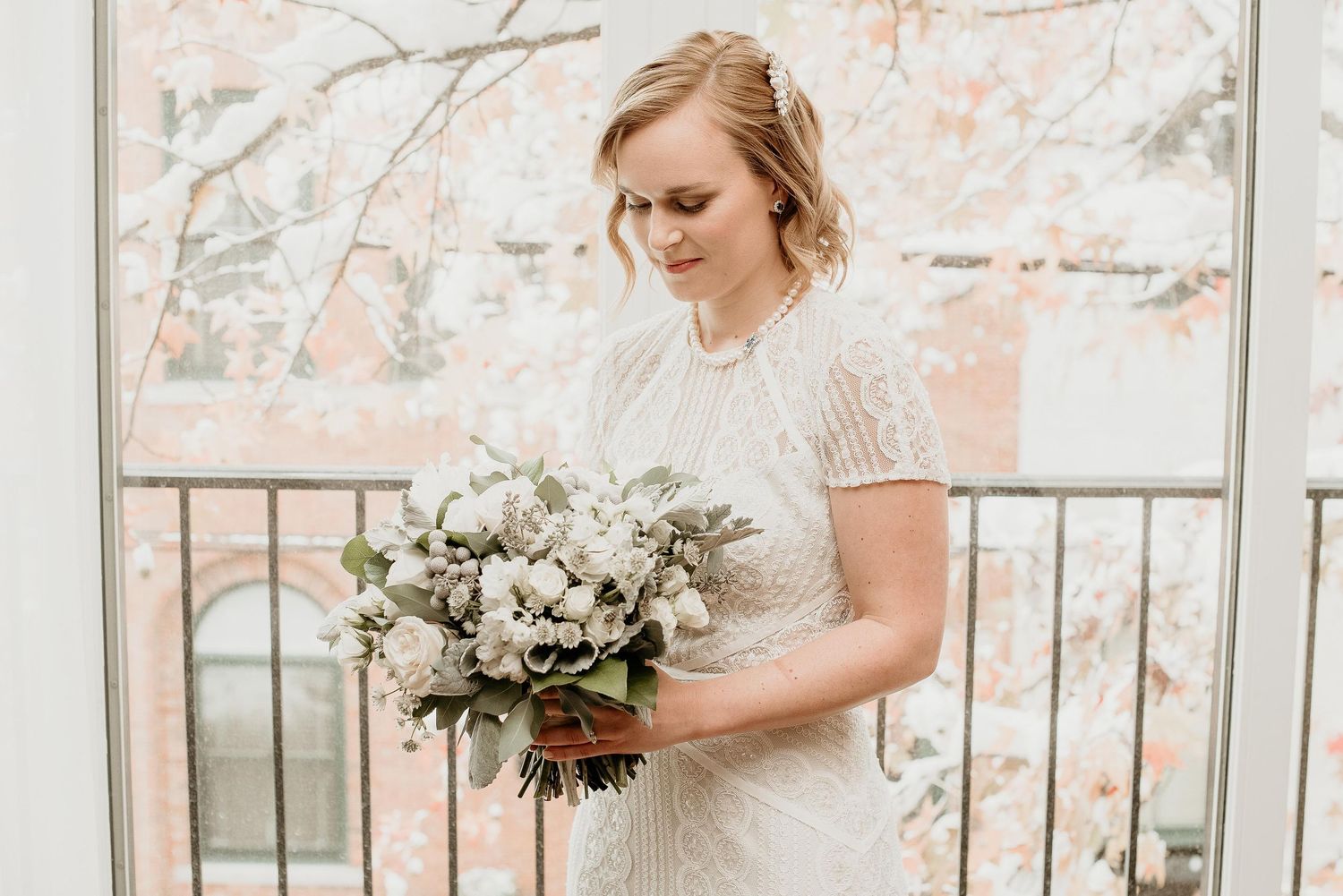 Bride in beaded white gown holding lush white and green bouquet, standing on balcony with autumn trees in background.