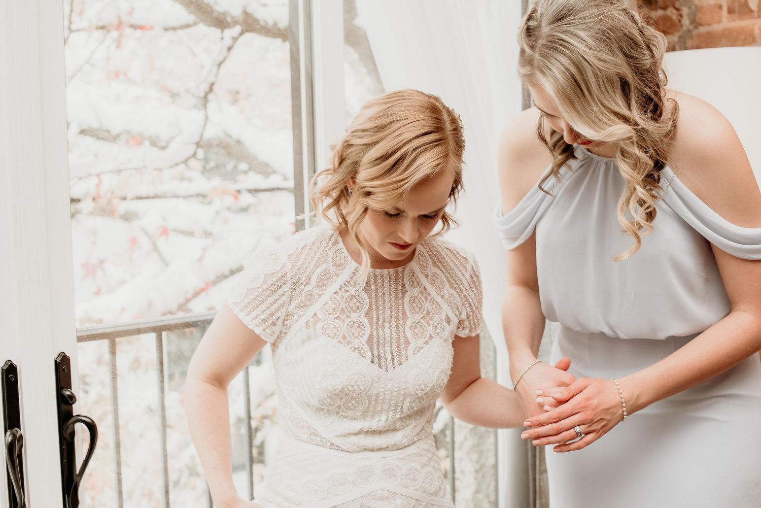 Bride in lace wedding dress being helped by bridesmaid in gray off-shoulder dress near bright window.