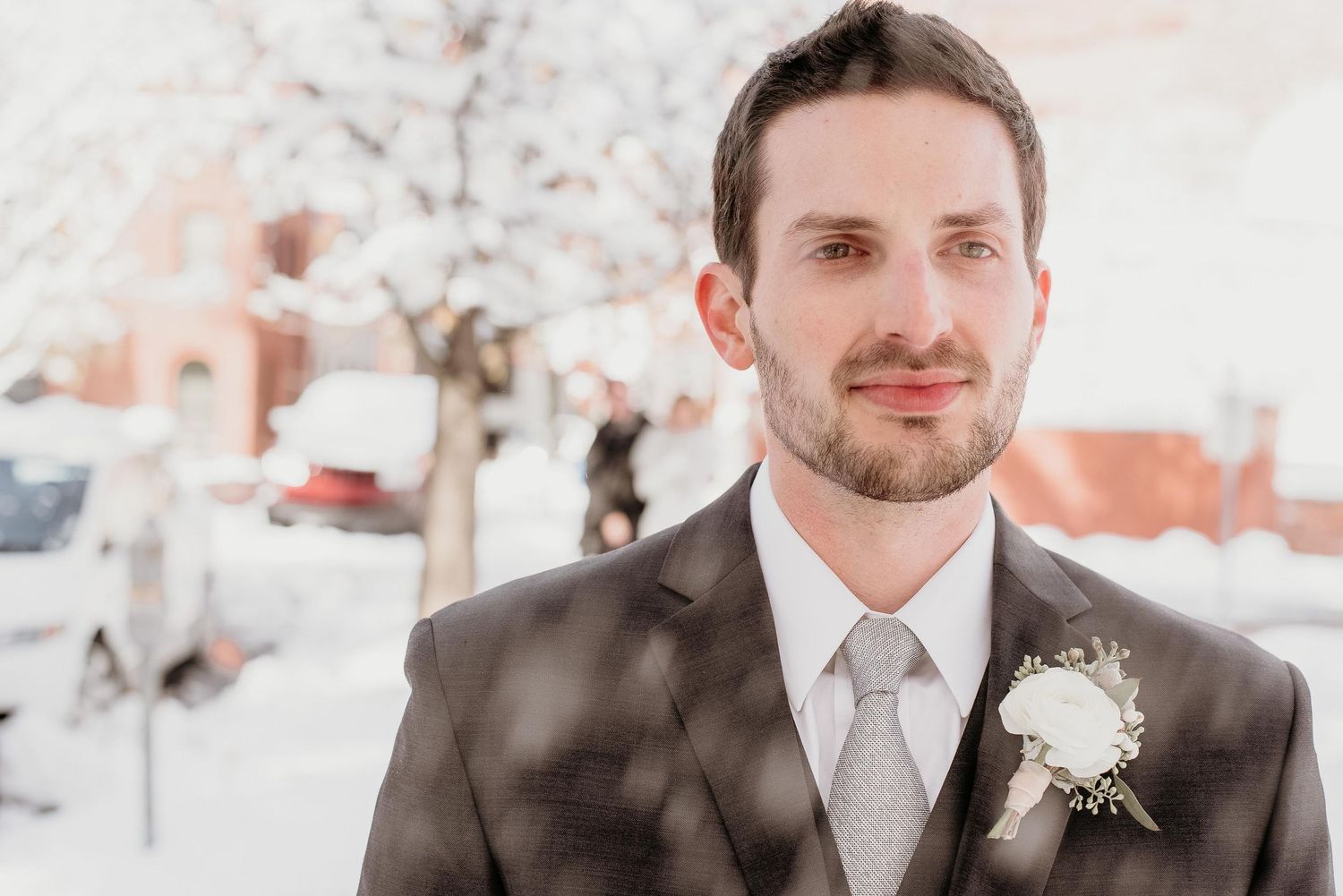 A groom in a gray suit with a white boutonniere stands outdoors in a snowy winter setting on his wedding day.