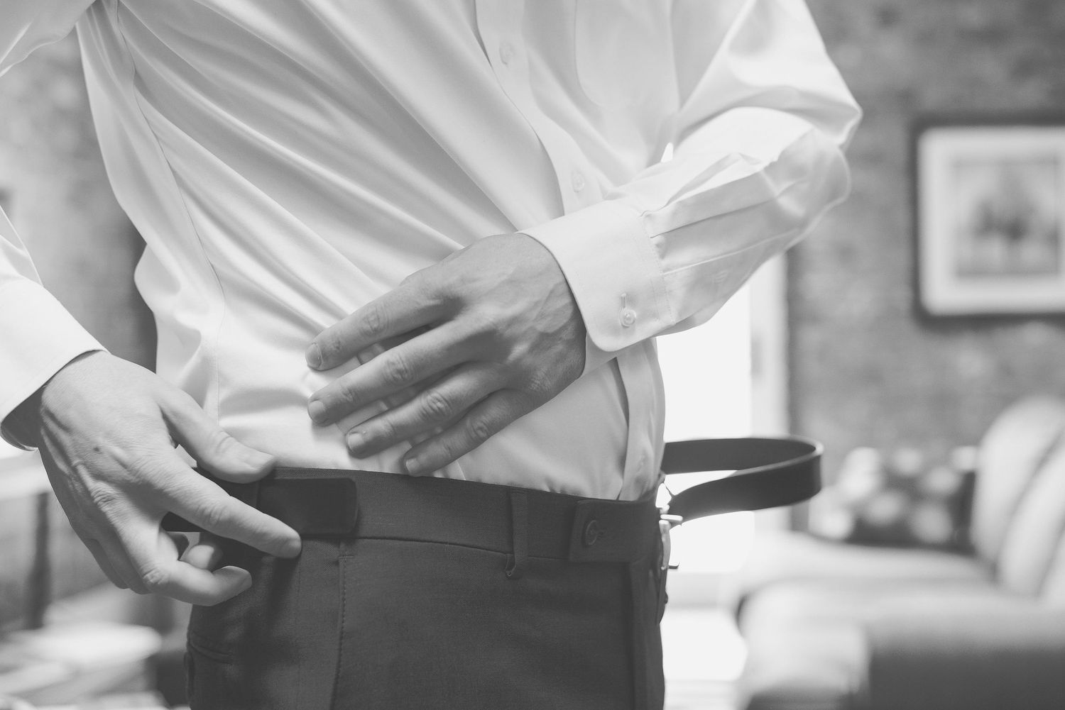Black and white photo of a man in a white dress shirt fastening a leather belt on dark trousers.