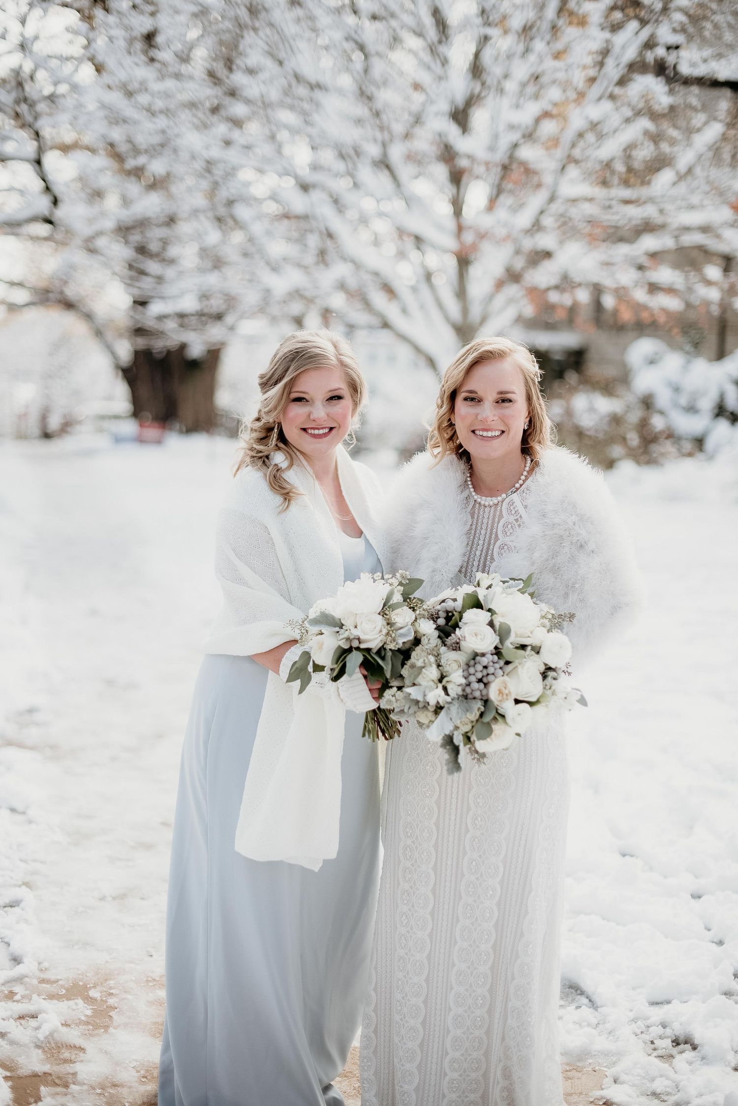 Two brides in winter wedding attire pose outdoors in snowy setting, one holding white floral bouquet.