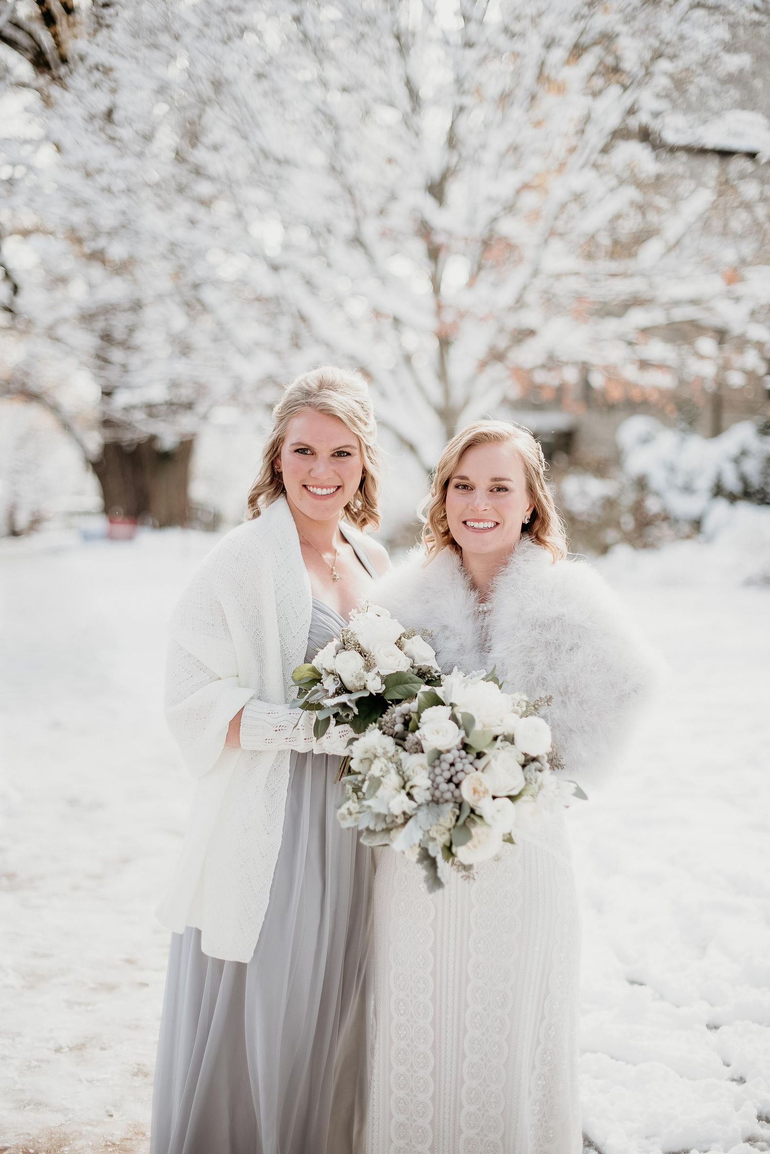 Two brides in white fur wraps hold a bouquet of white flowers while posing in a snowy winter wedding outdoor setting.
