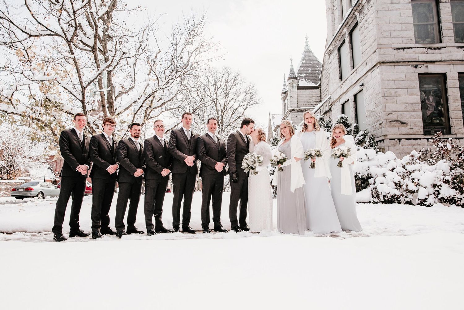 Wedding party in dark suits and gray dresses posing in snow outside a historic building in winter.