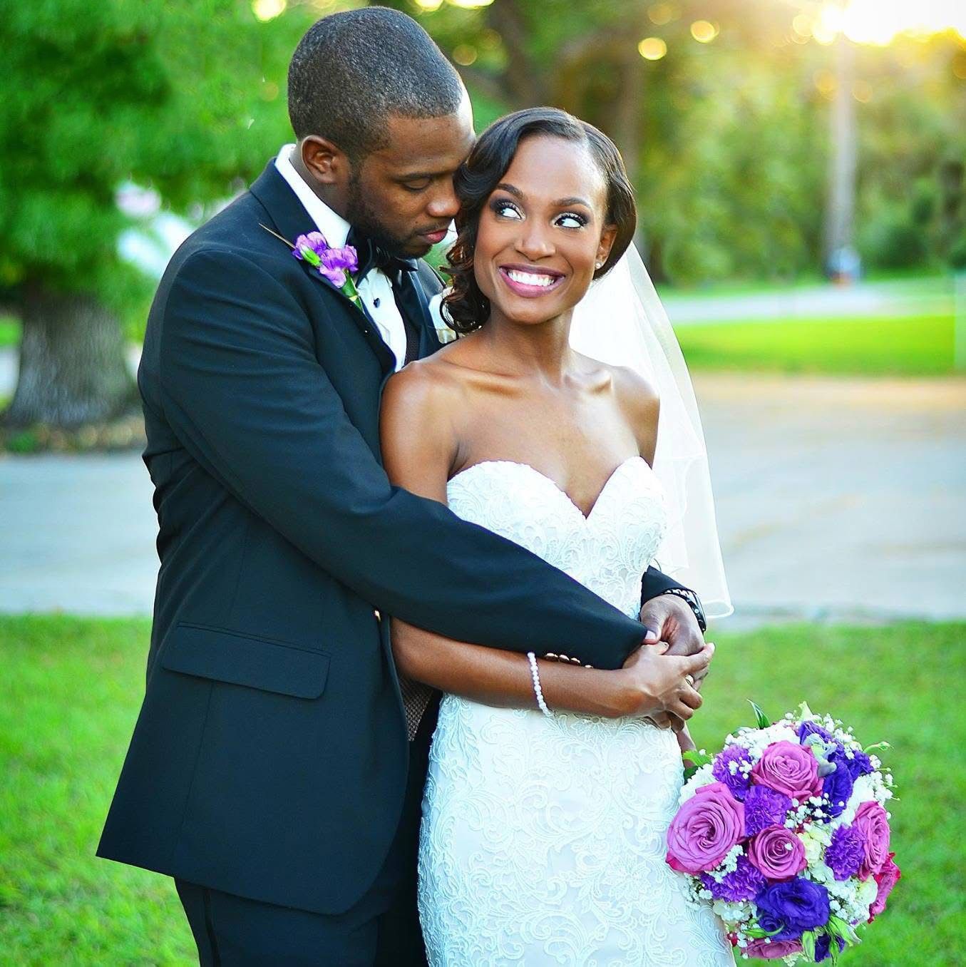 Elegant bride in lace gown holding purple rose bouquet with groom in tuxedo.