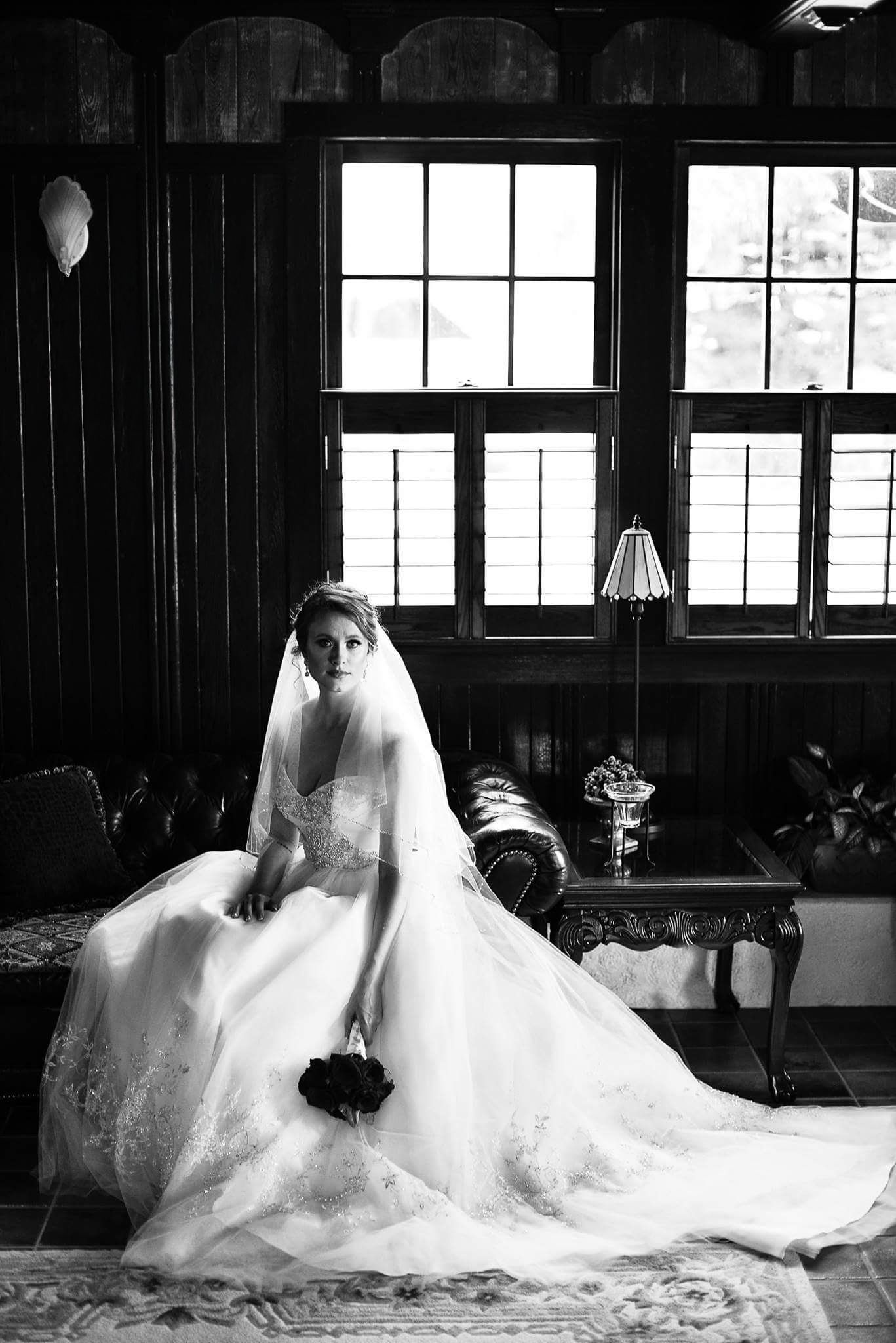 Black and white portrait of a bride in a full lace gown seated near dark wood windows holding roses.