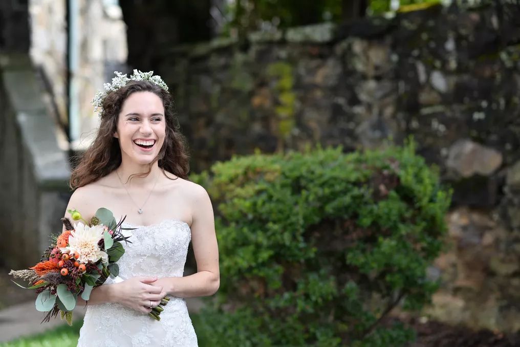 Smiling bride in strapless lace gown holds autumn bouquet wearing floral crown near stone wall outdoors.