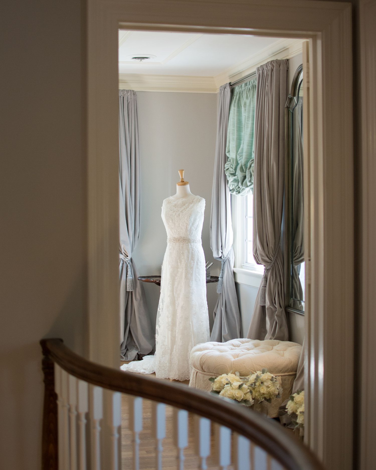 Elegant lace wedding dress displayed on mannequin in softly lit bridal suite with gray curtains.