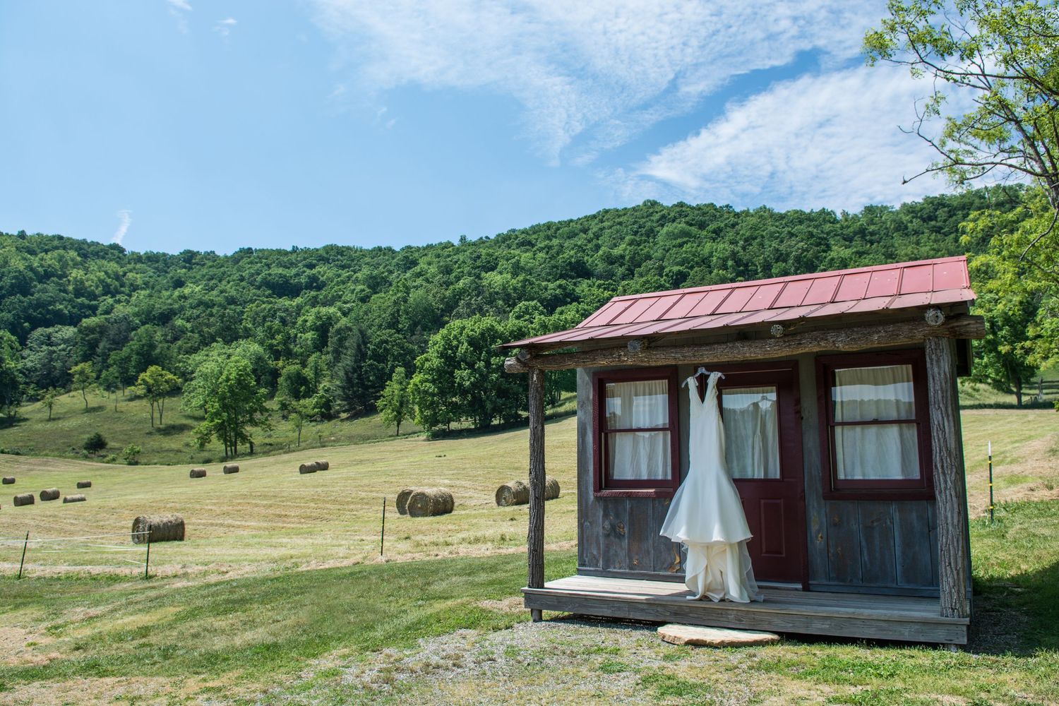 White wedding dress hanging on rustic red-roofed cabin with scenic countryside hay bales and green hills.