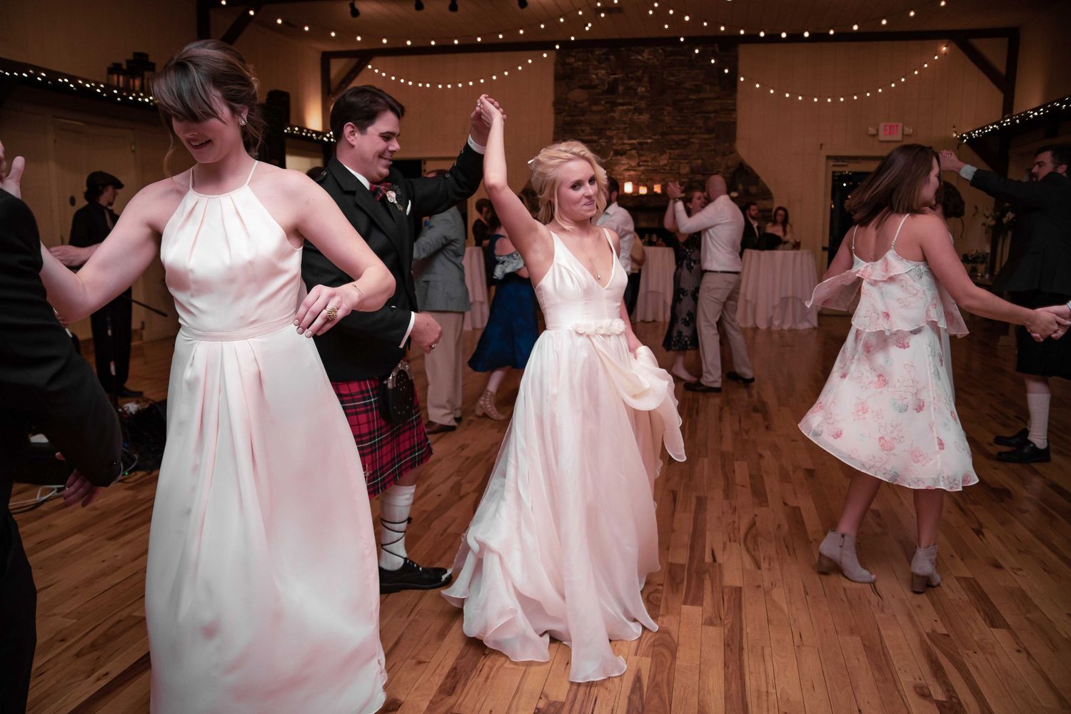Women in elegant white gowns dancing joyfully at a wedding reception on a wooden dance floor with warm string lights.