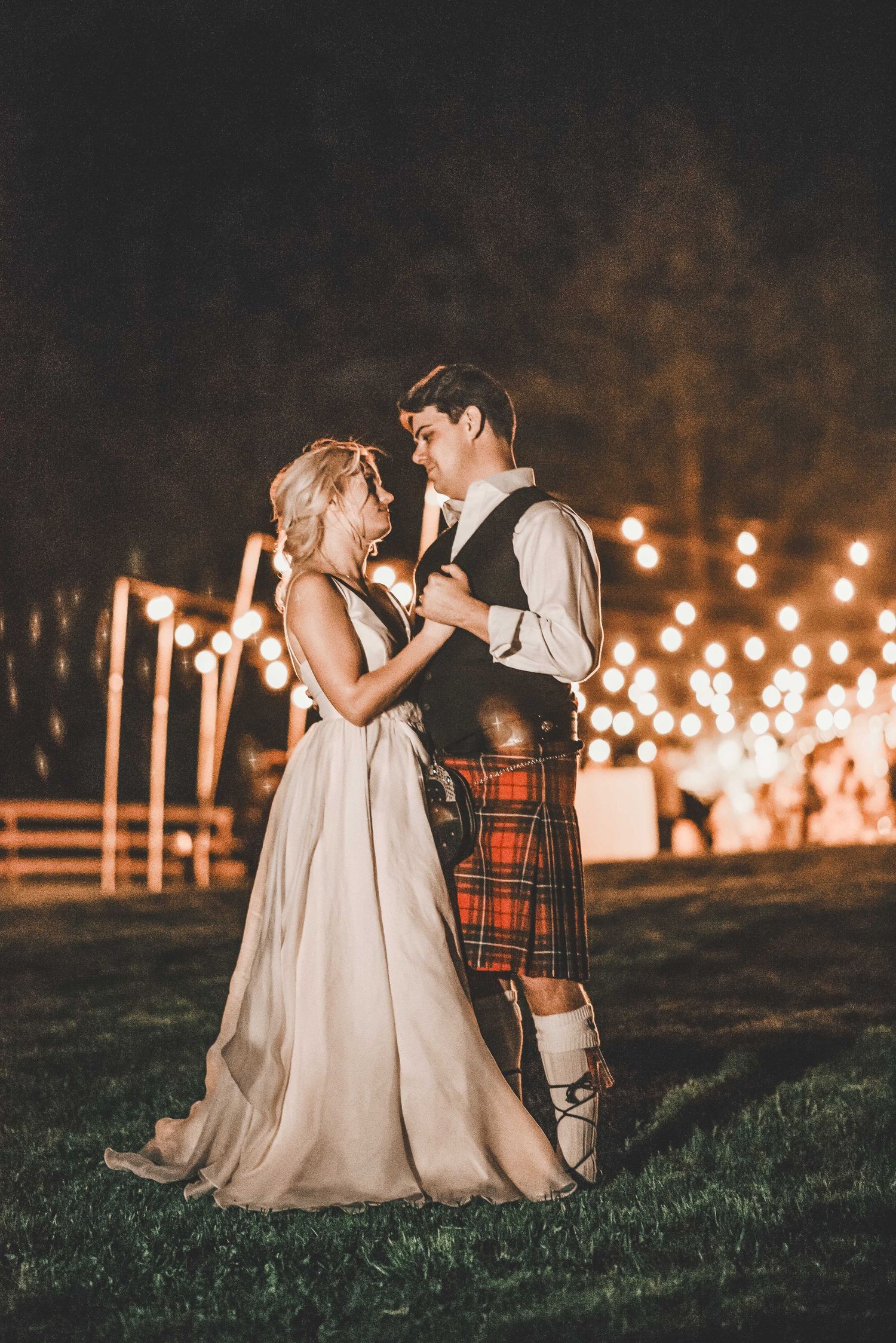 Bride and groom share first dance outdoors at night, groom wearing traditional Scottish kilt, surrounded by warm string lights.