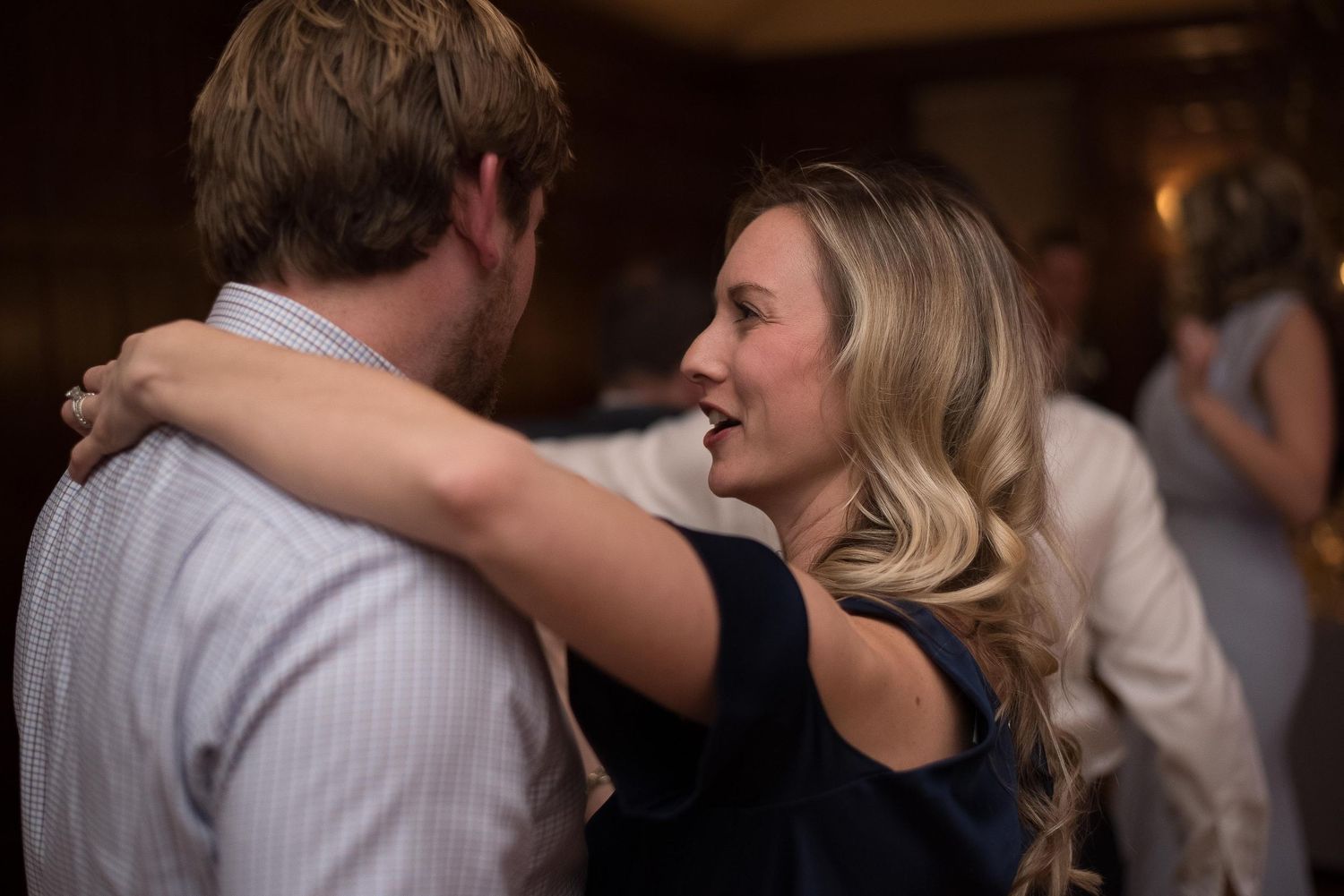 A smiling blonde woman in a navy dress dancing with a man in a striped shirt at a social event.