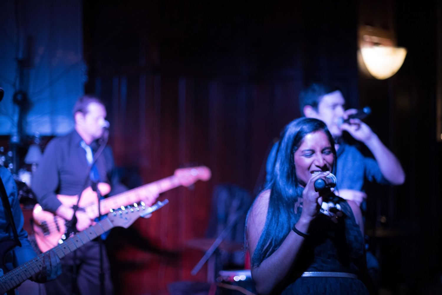 Live band performance with female vocalist singing into mic and guitarist playing red electric guitar on dimly lit stage.