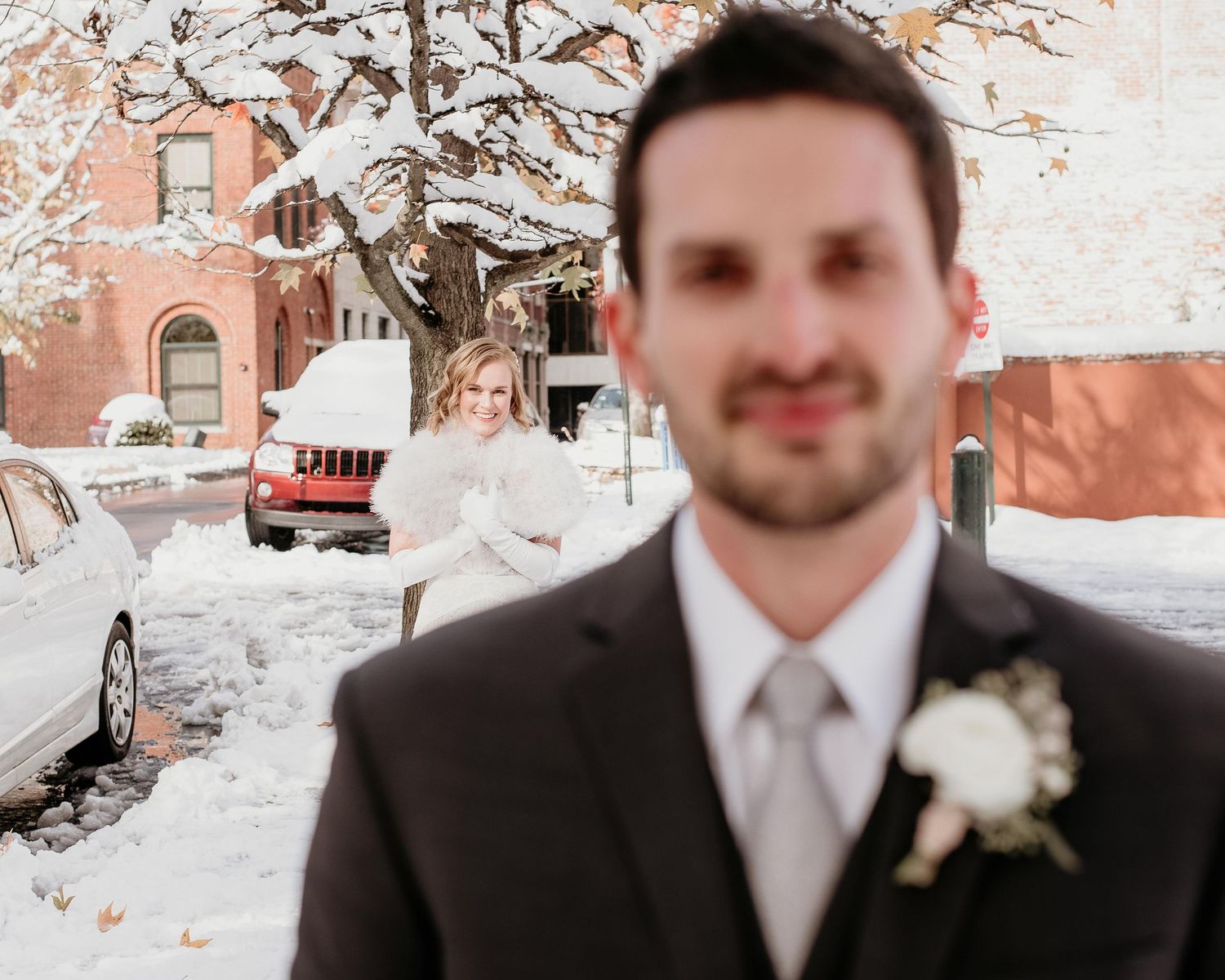 Groom in gray tie with boutonniere stands in foreground as bride smiles in snowy winter wedding outdoor scene.