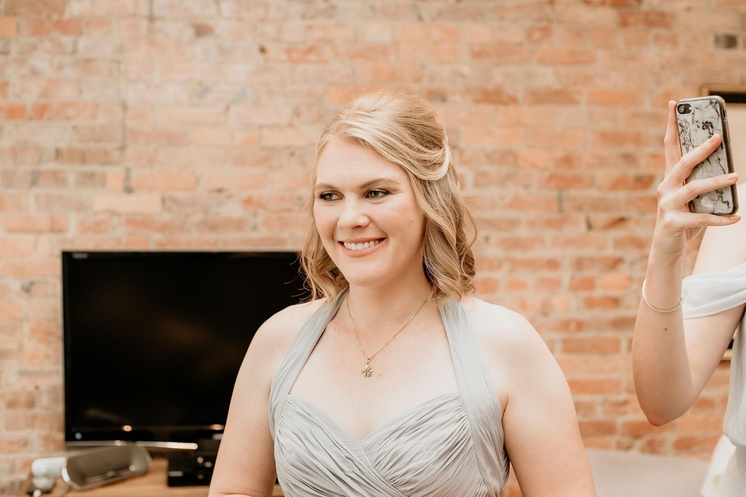 Smiling bridesmaid in silver dress taking selfie against rustic brick wall backdrop.