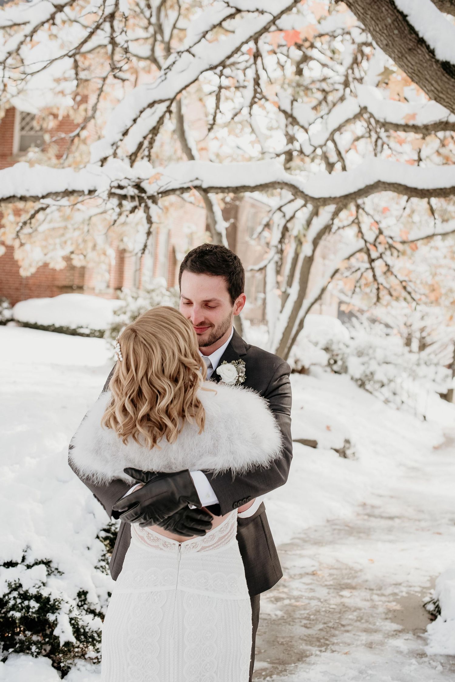 Bride and groom embrace in snowy winter garden, bride wearing white gown with fur stole, groom in dark suit with gloves.