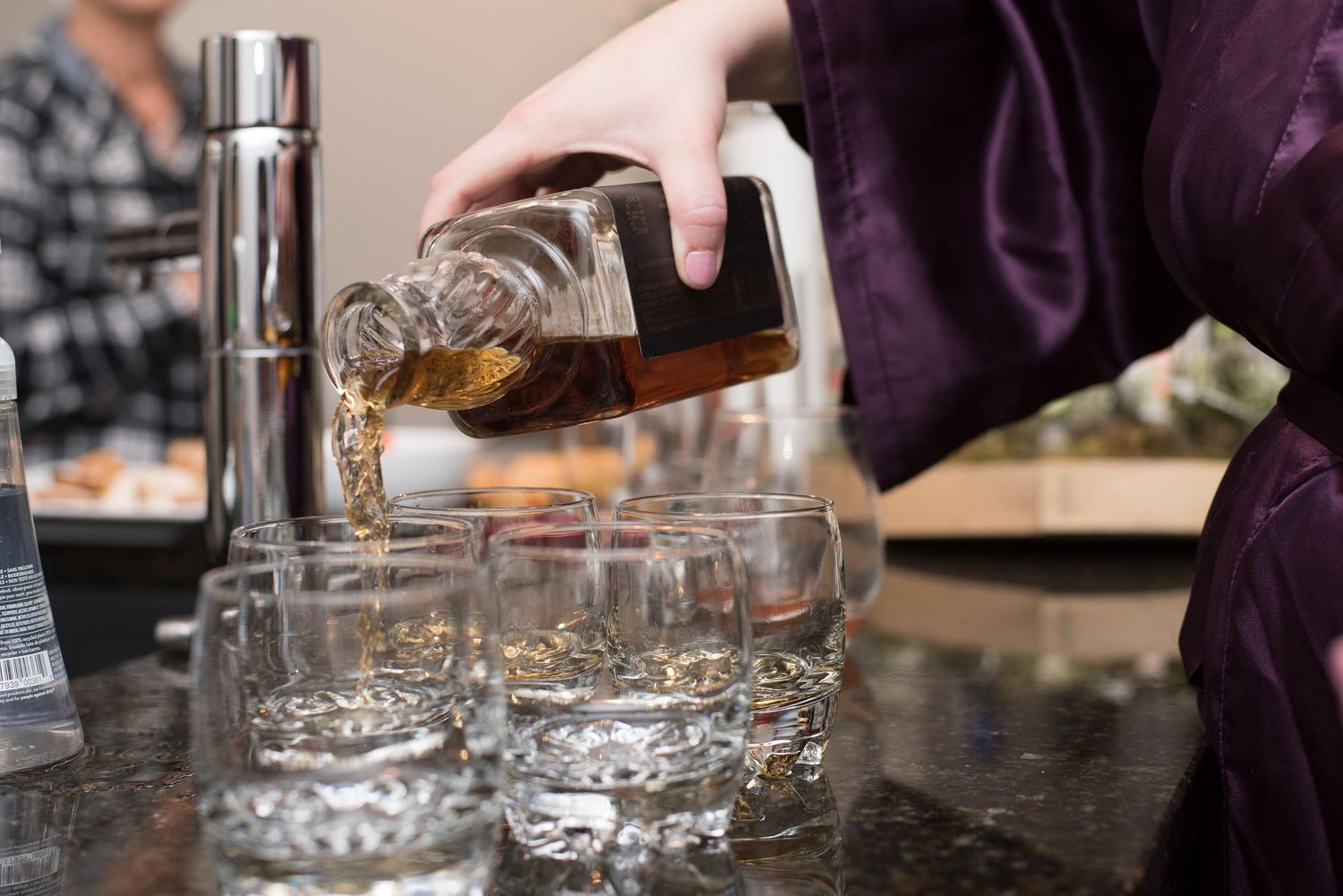 Bartender pouring amber whiskey from a bottle into multiple crystal shot glasses on a bar counter.