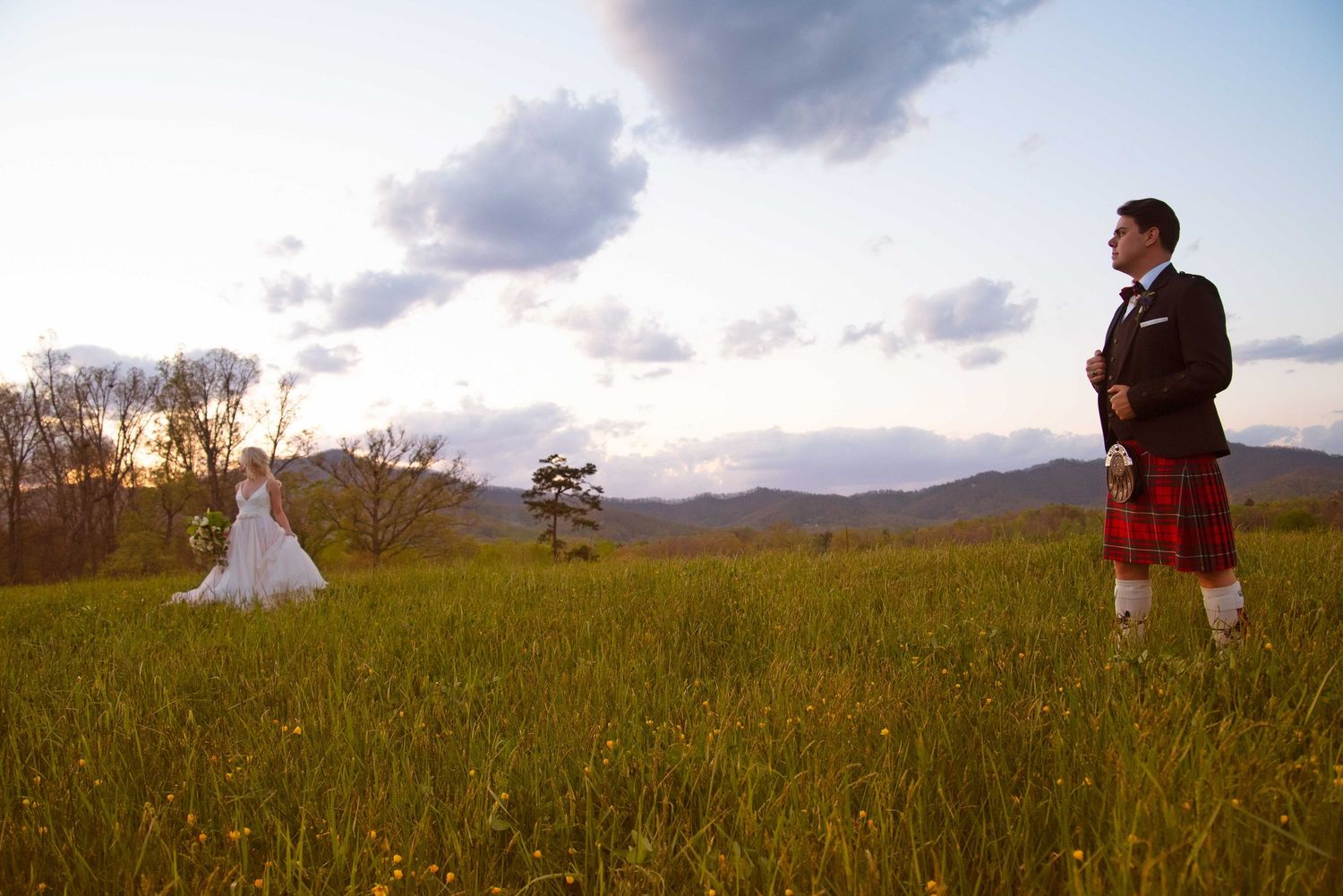 Groom in red tartan kilt stands in green meadow with bride in white gown, mountains and sunset sky behind them.