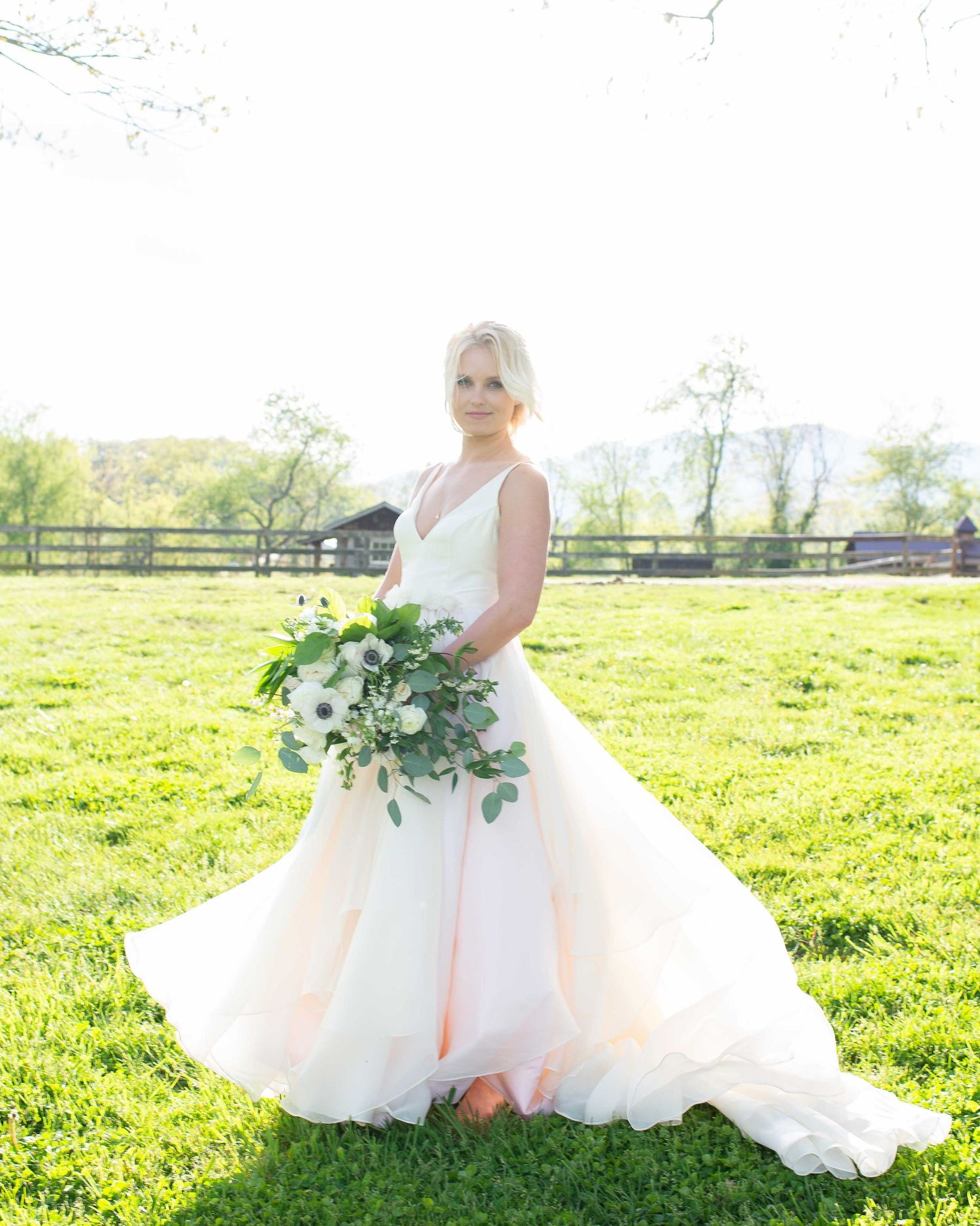 Blonde bride in blush pink ballgown holding white floral bouquet standing in green field at outdoor farm wedding.
