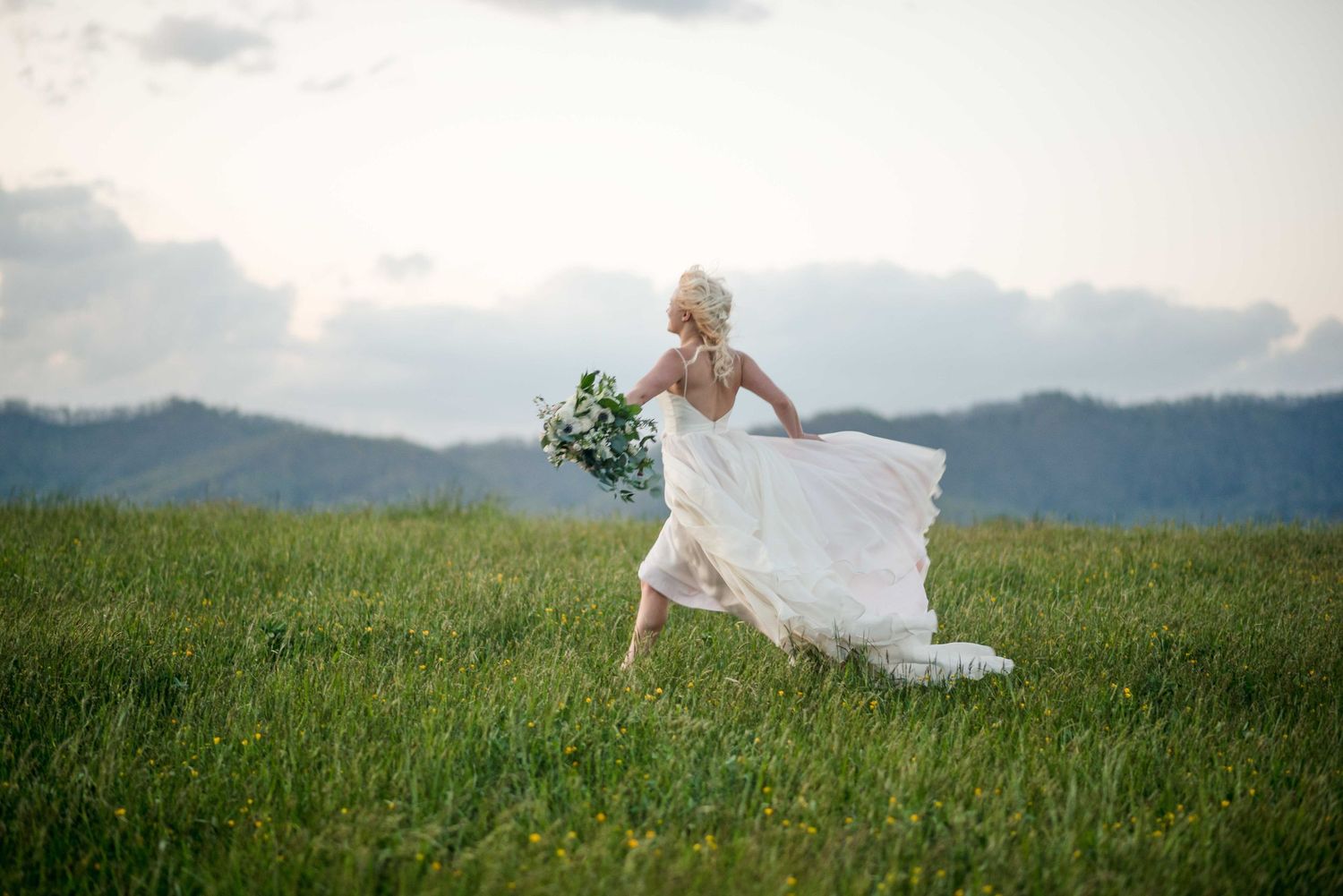 Bride in white gown running through lush green meadow with mountain backdrop, holding bouquet.