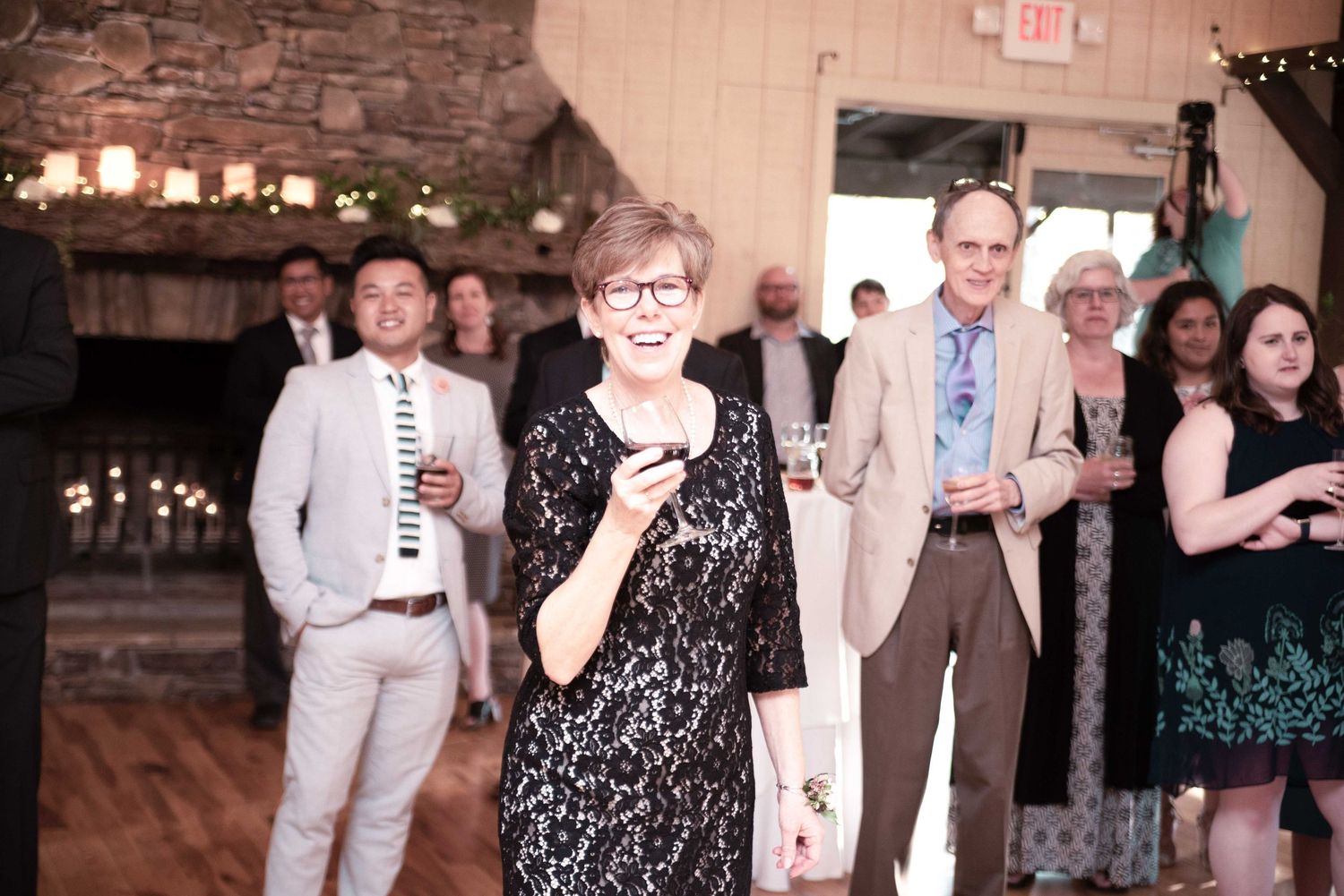 Woman in black lace dress laughing at indoor wedding reception with guests socializing in background.