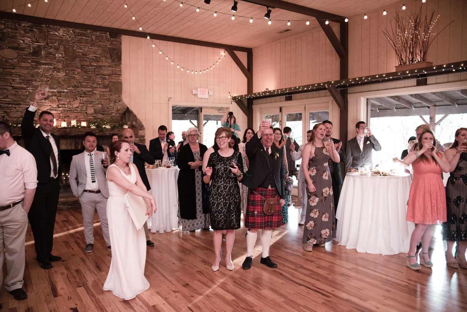 Bride tosses bouquet to excited wedding guests on dance floor in rustic barn venue with string lights.