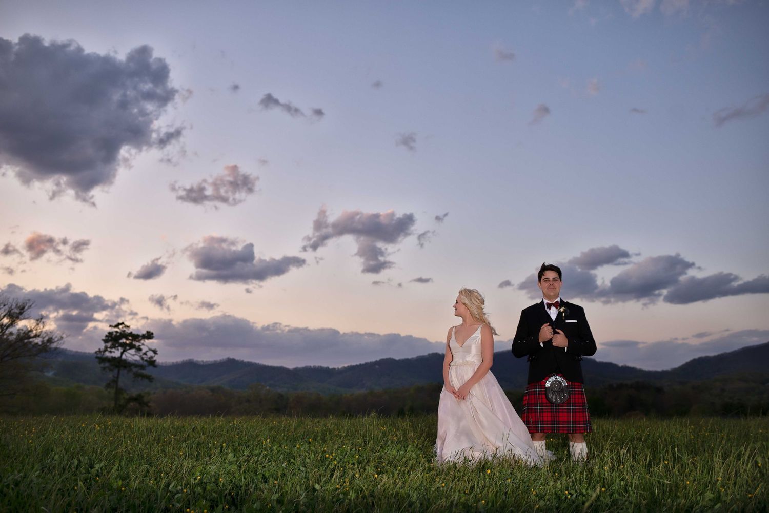 Bride in pink gown and groom in tartan kilt stand in green field at sunset with mountain backdrop.